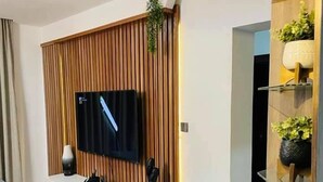 A modern living room with a flat-screen TV mounted on a wooden panel wall, a white entertainment unit below, and a black countertop. - Winnie Host and Homes (Abuja)