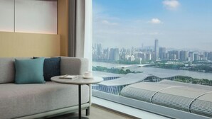 A modern hotel room with a large window offering a cityscape view, a sofa with cushions, and a small table with a book and a cup. - Courtyard By Marriott Guangzhou Pazhou (Guangzhou)