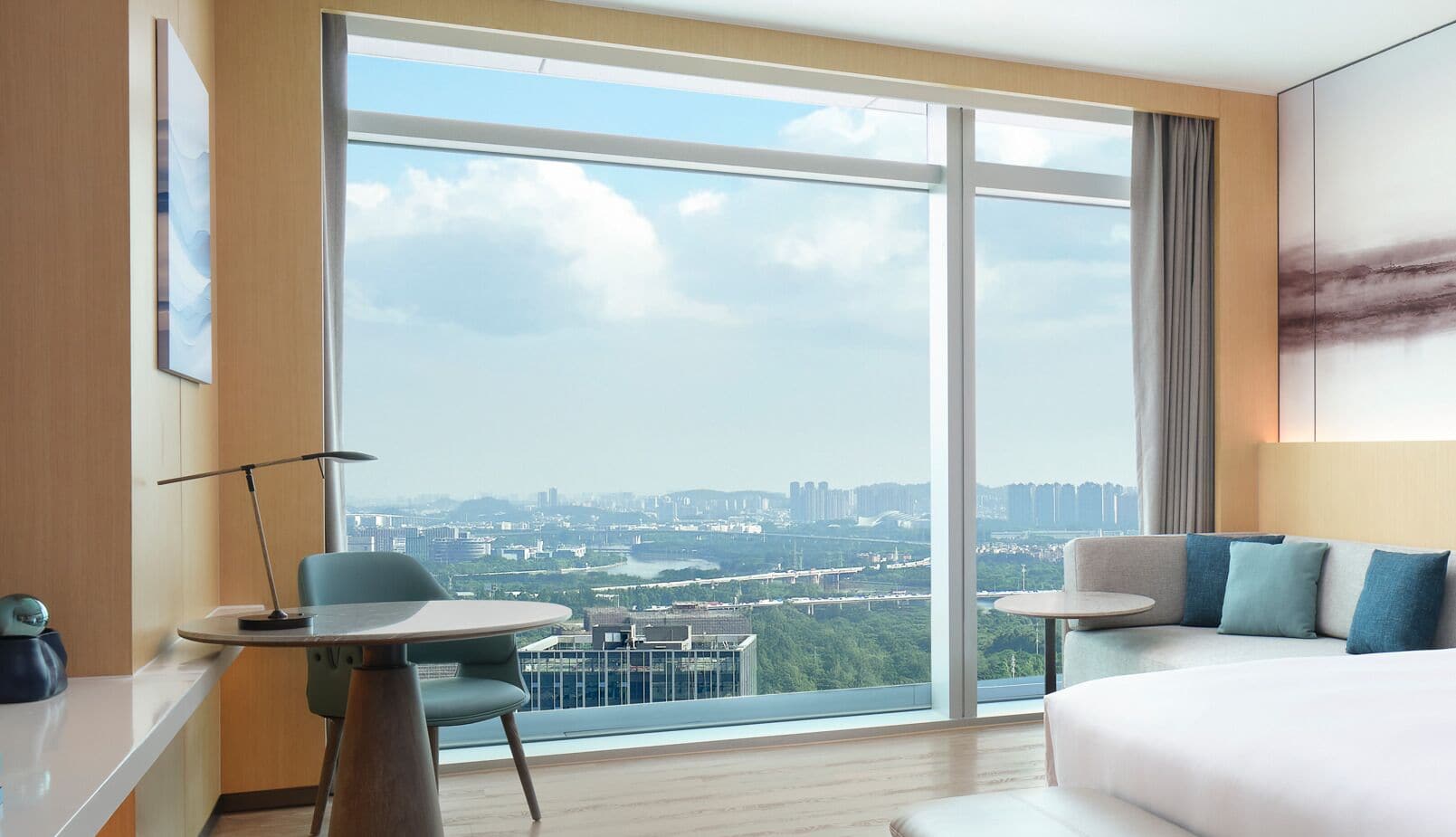 A hotel room with a large window, a bed, a sofa, a small table, and a chair.
