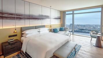 Una habitación de hotel moderna con una cama grande, mesitas de noche, un escritorio y vistas a un paisaje urbano a través de amplios ventanales.