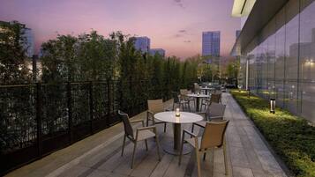 An outdoor seating area with tables and chairs, a glass building to the right, and a city skyline in the background.