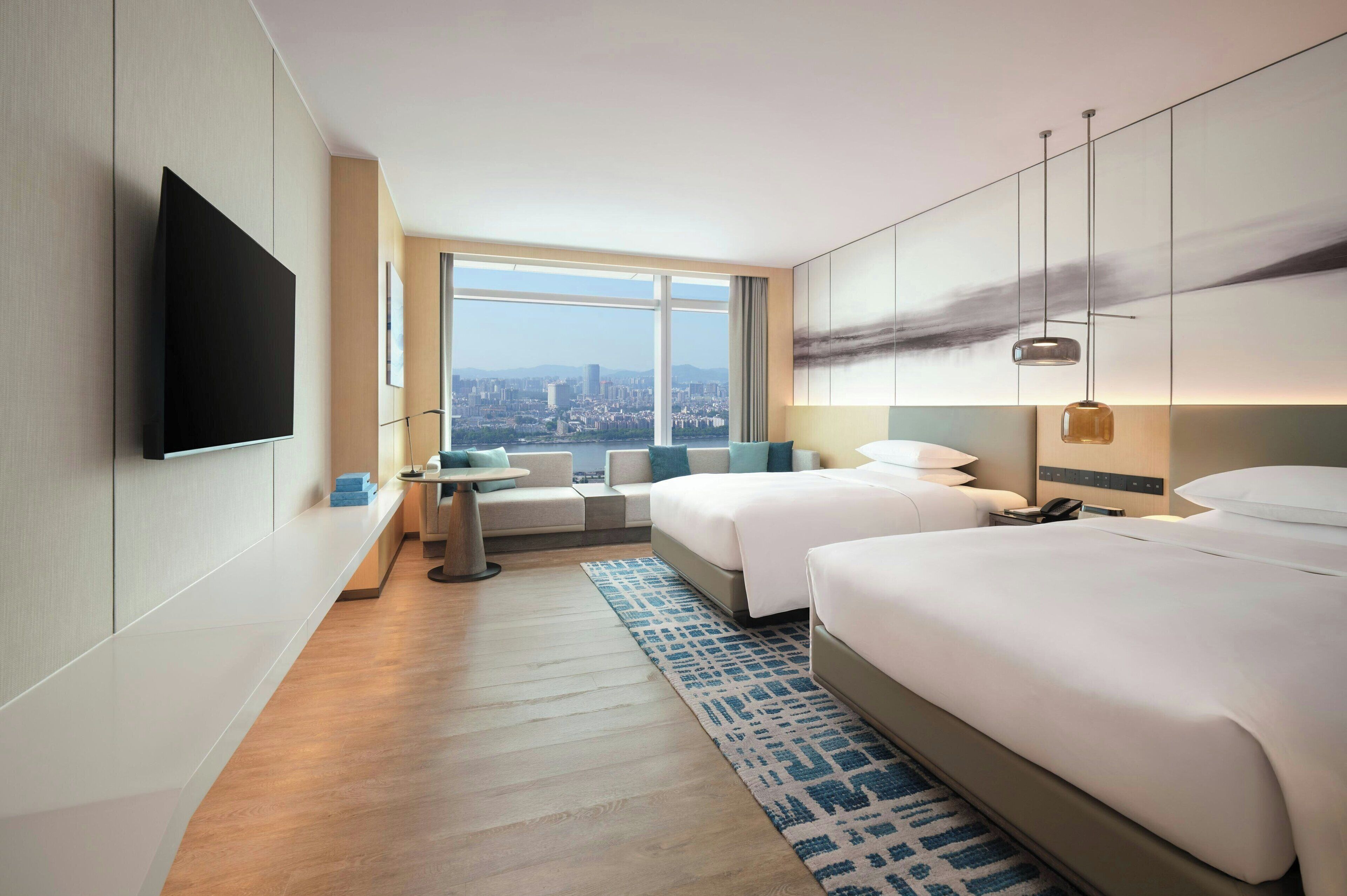 A hotel room with two beds, a TV, a city view, and a small seating area.