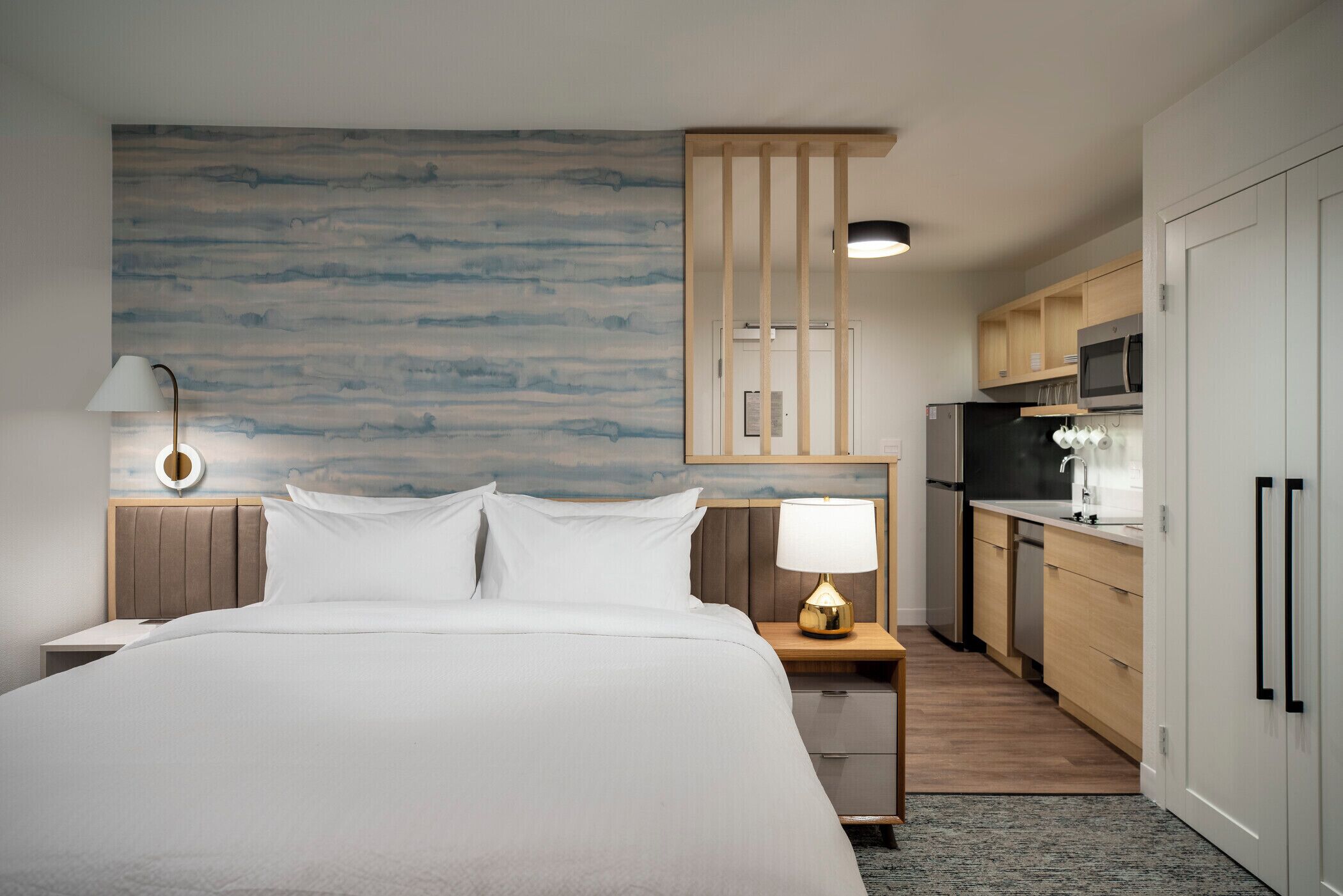 A hotel room with a bed, bedside lamp, nightstand, wooden headboard, and a view of the kitchen area.