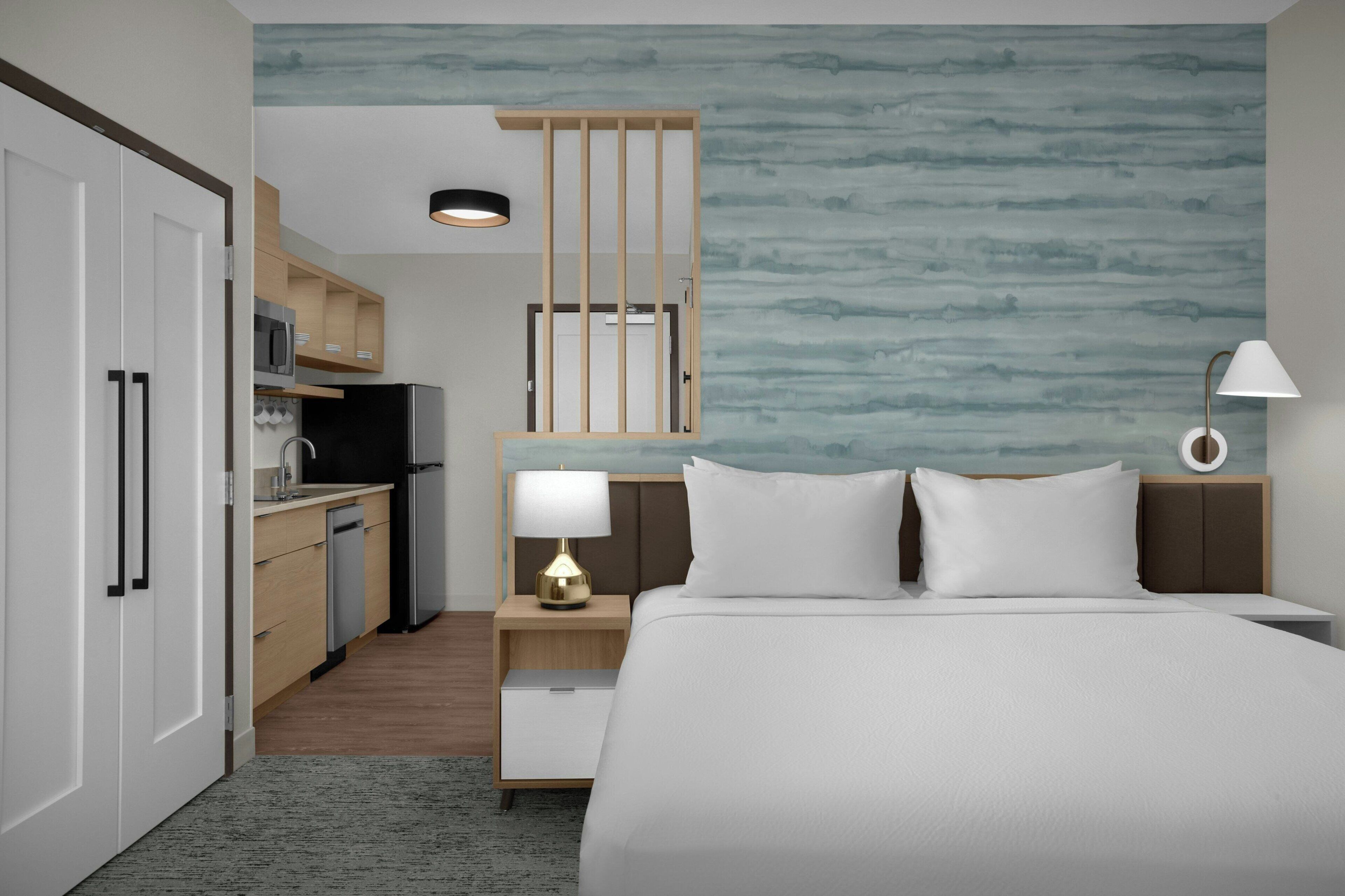 A hotel room with a bed, bedside tables, a desk, a kitchenette, and a bathroom.