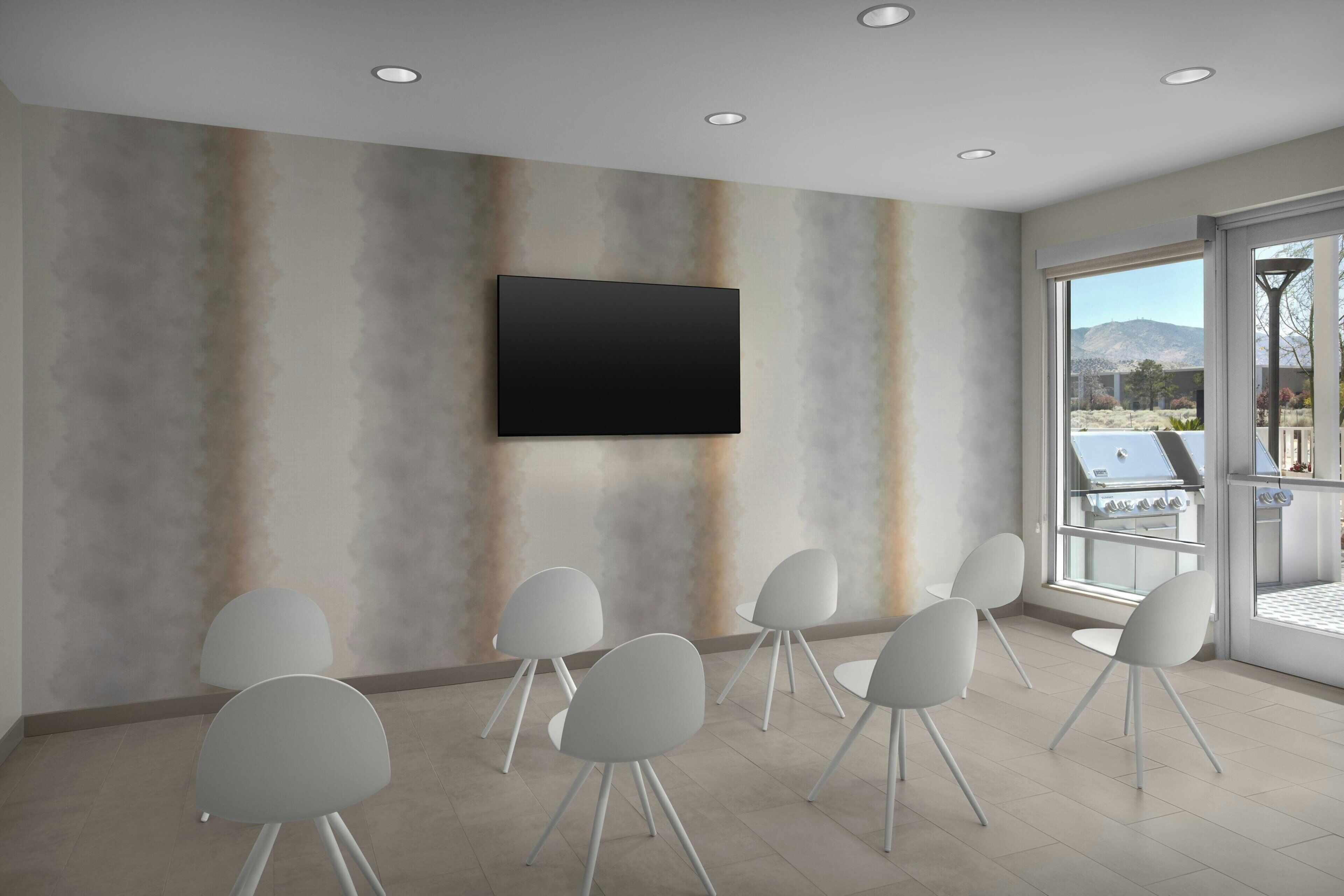 A conference room with a flat-screen TV mounted on the wall, surrounded by white chairs with metal legs.