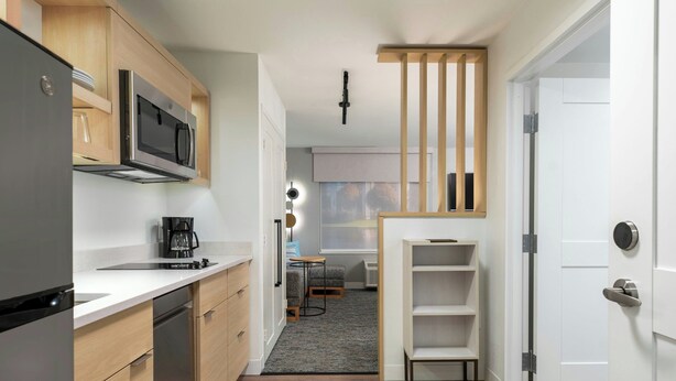 A modern kitchen with a refrigerator, microwave, and coffee maker. There is a small dining area with a table and chairs. A wooden shelf unit is visible, and a door with a handle is on the right. - Towneplace Suites By Marriott Palmdale (Palmdale)