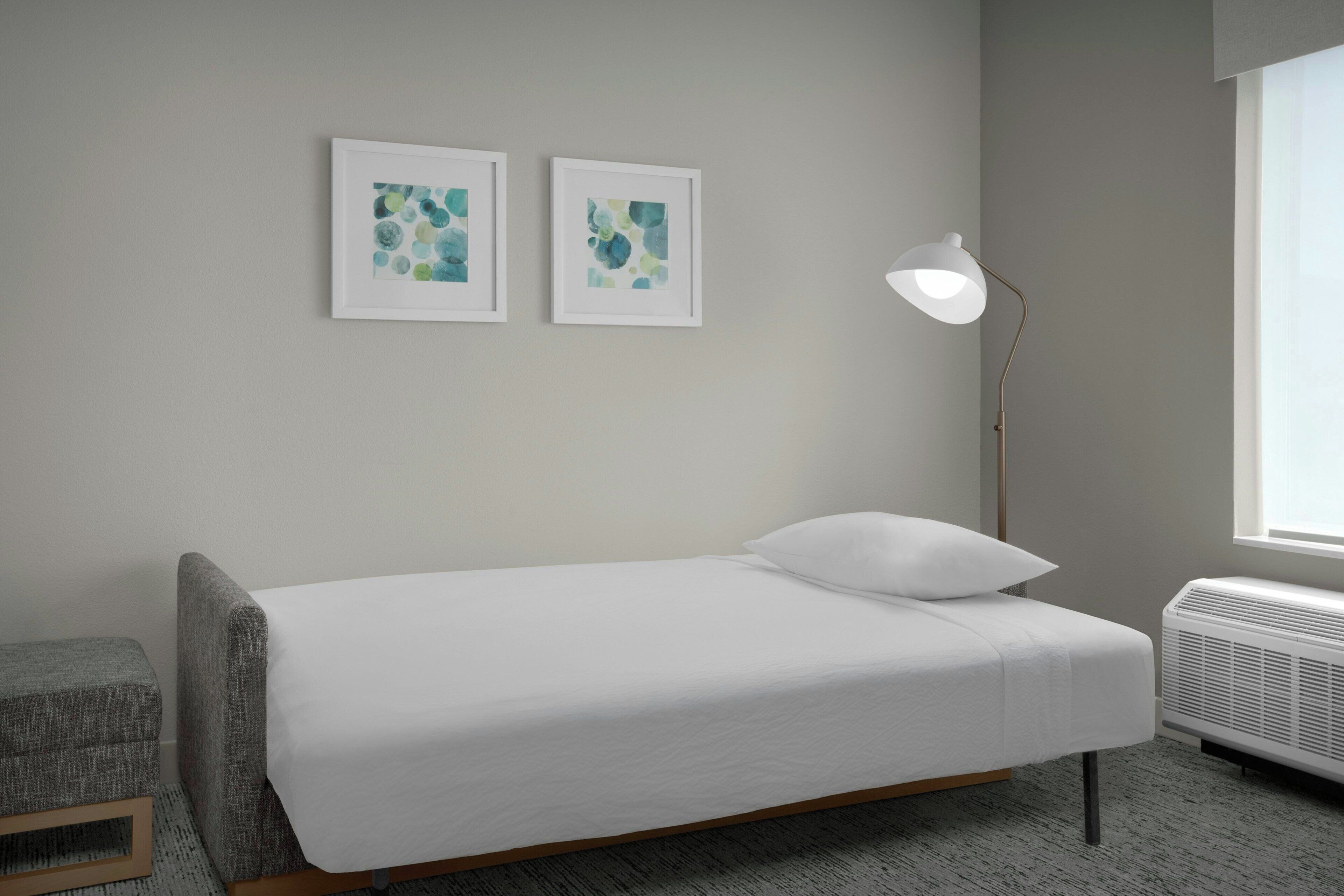 A single bed with a white mattress and pillow, a gray upholstered bench, a floor lamp, and two framed abstract paintings on the wall.