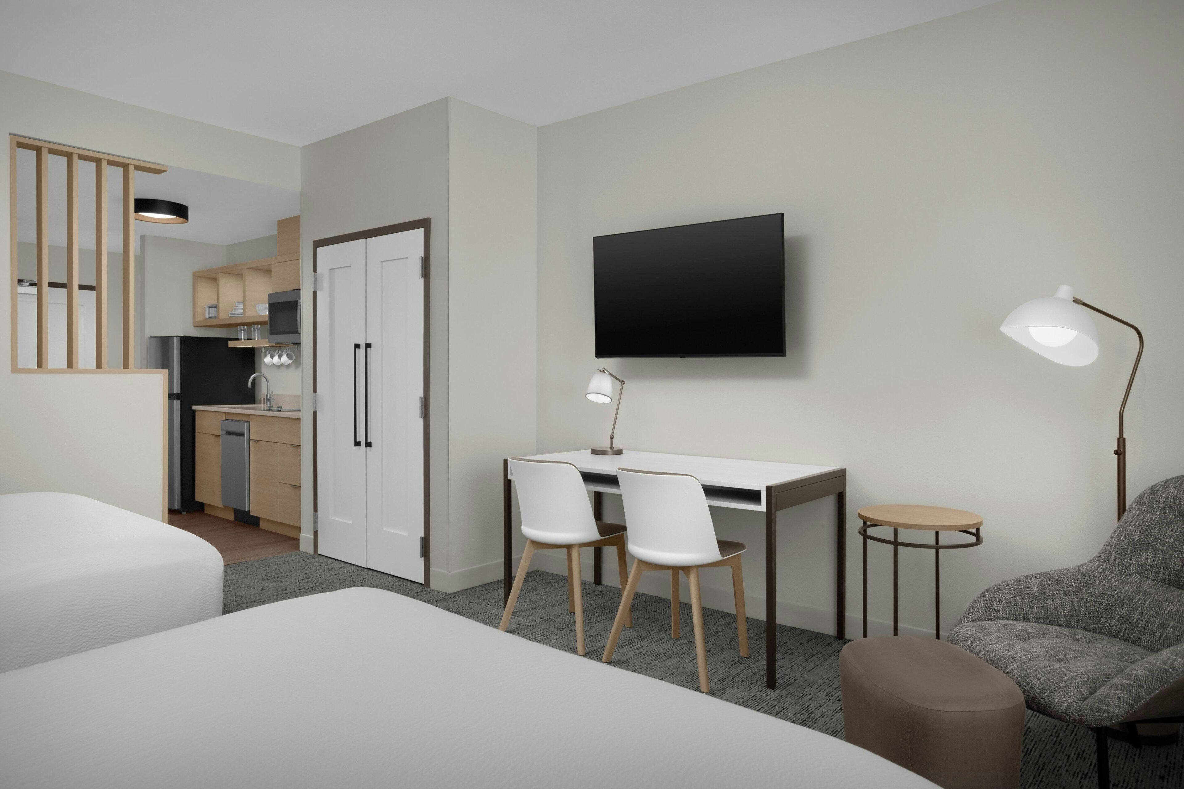A hotel room with a bed, a desk with two chairs, a TV, a lamp, and a kitchenette in the background.