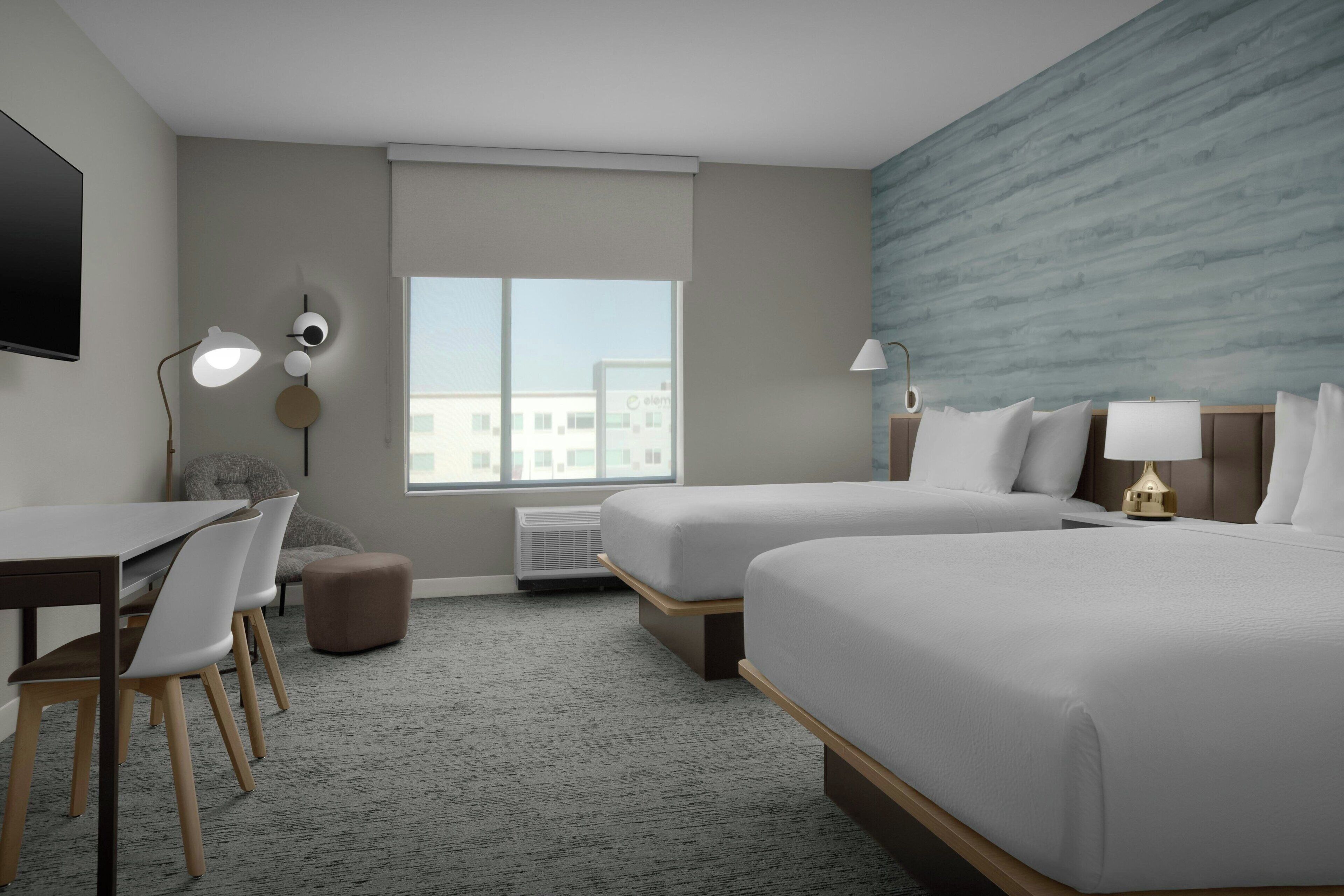 A hotel room with two beds, a desk, chairs, a TV, and a large window.