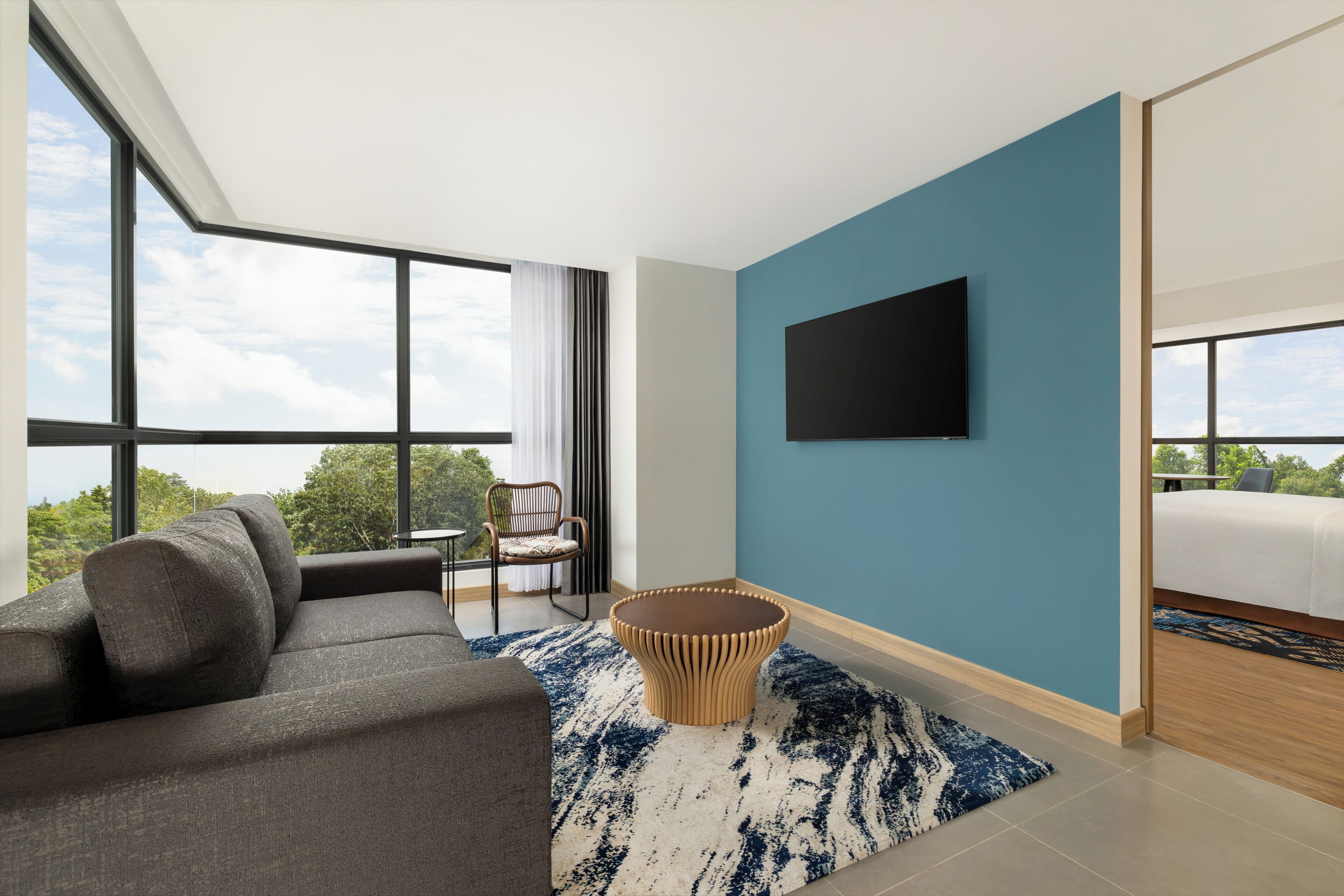 A modern hotel room with a large window, a flat-screen TV, a sofa, a round coffee table, and a bed.