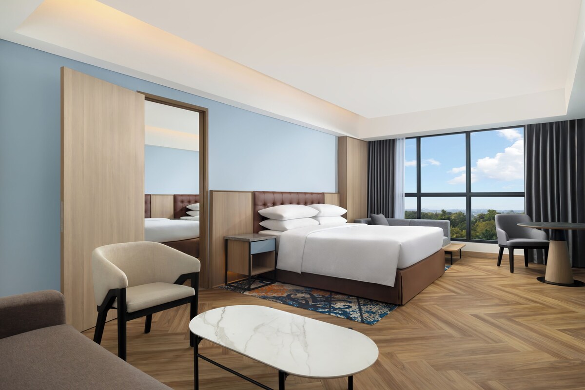 A modern hotel room with a large bed, a desk, a chair, and a view of the outdoors.