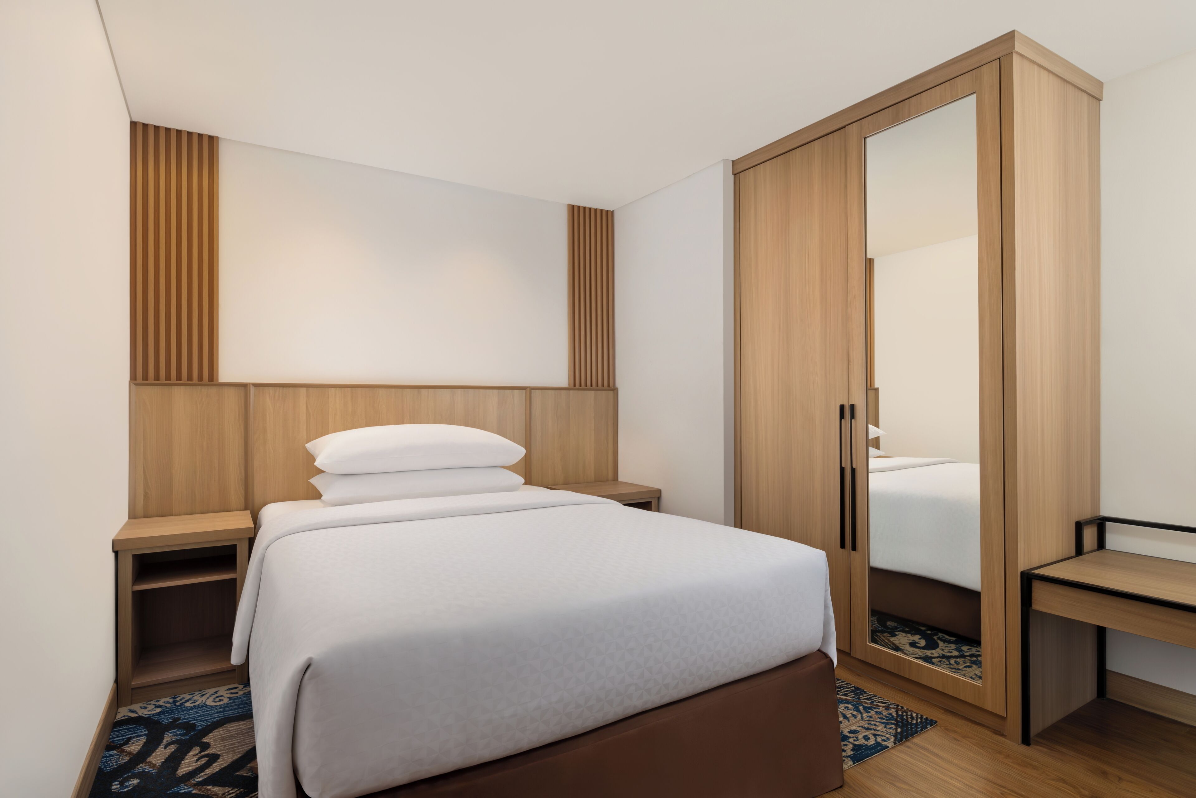 A modern hotel room with a bed, a wooden wardrobe, a bedside table, and a mirror.