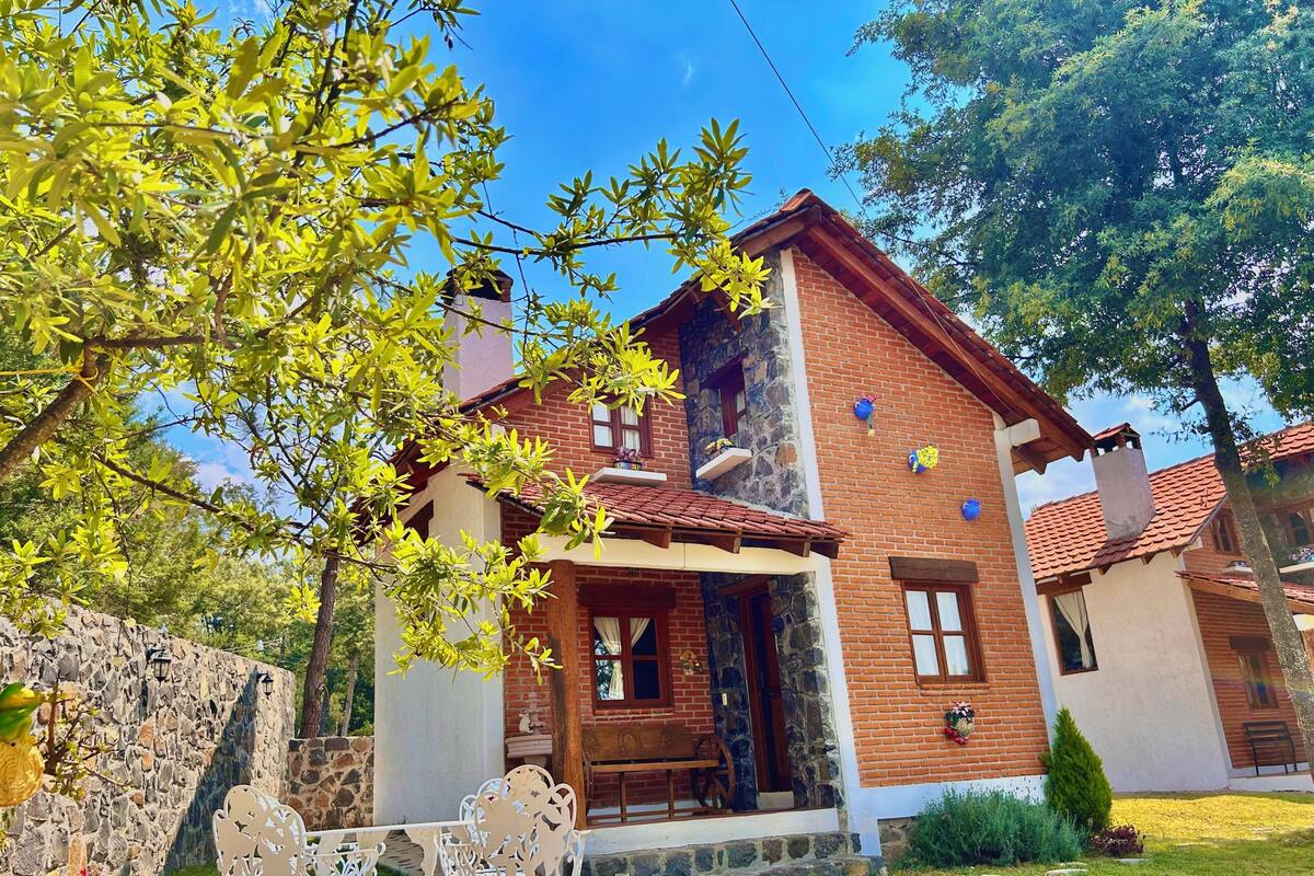 A two-story house with a stone wall, a wooden bench, and a table in the front yard.