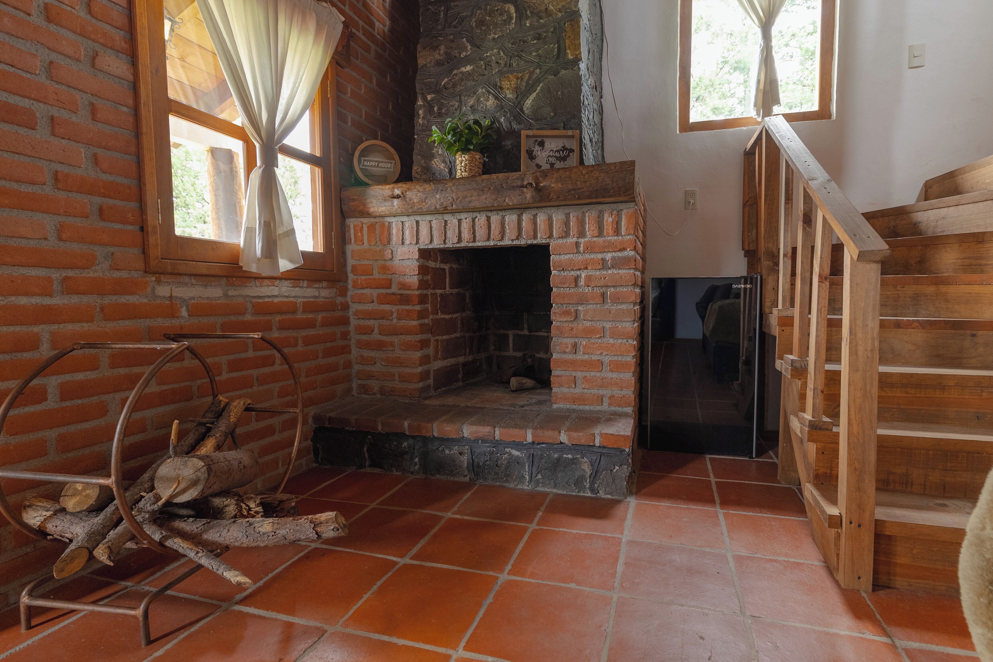 Una habitación rústica con chimenea de ladrillo, escalera de madera y una ventana con cortinas.