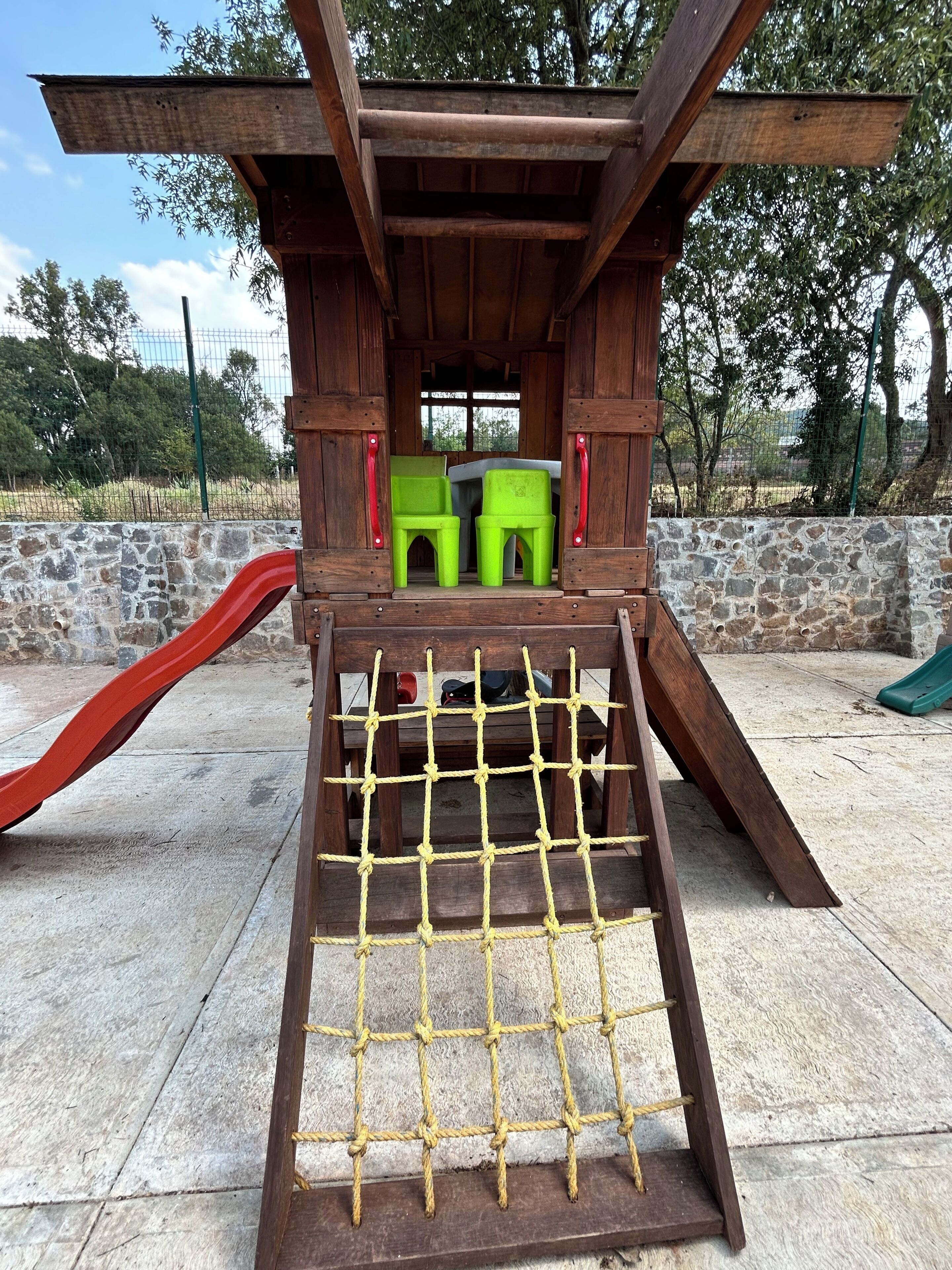 Una estructura de juego de madera con un tobogán, una escalera de cuerda y dos sillas verdes.
