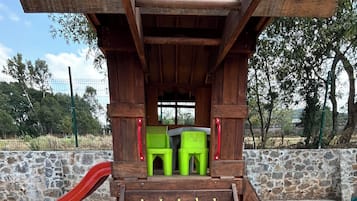 Una estructura de juego de madera con un tobogán, una escalera de cuerda y dos sillas verdes.