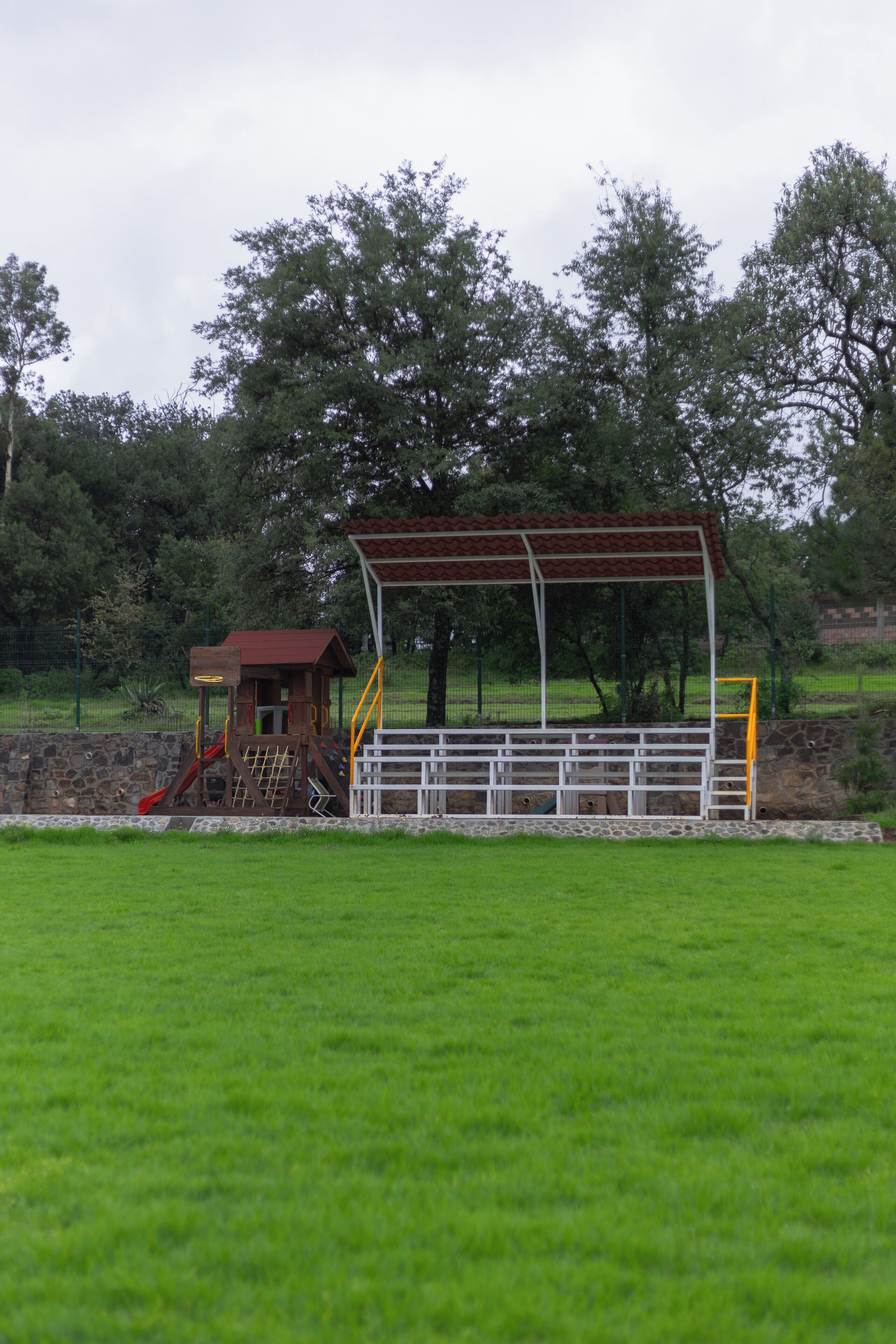 Un parque infantil con un tobogán y estructuras para escalar, rodeado de un muro de piedra y un campo de césped.