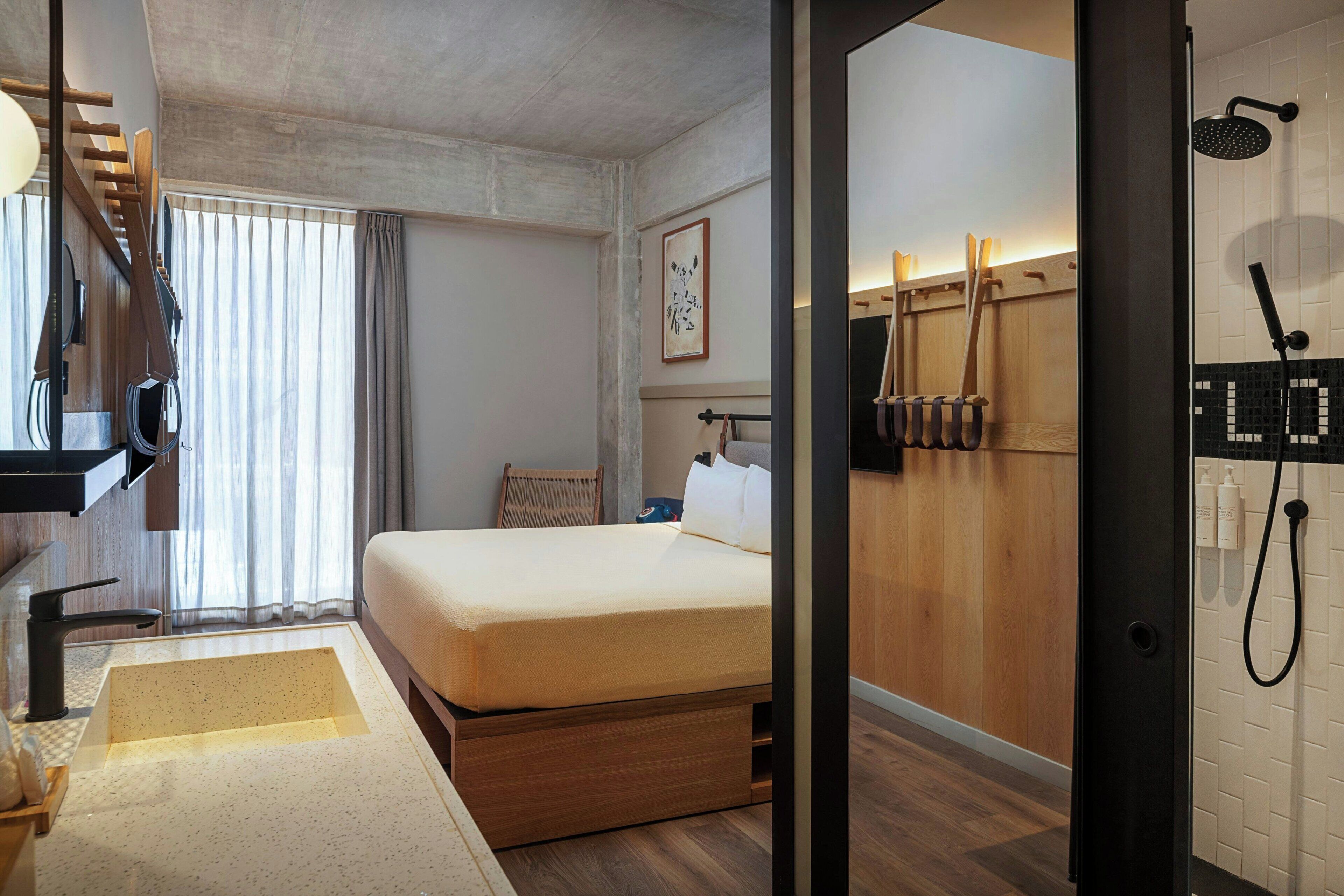 A hotel room with a bed, a bathroom with a shower, a wooden floor, and a window with curtains.