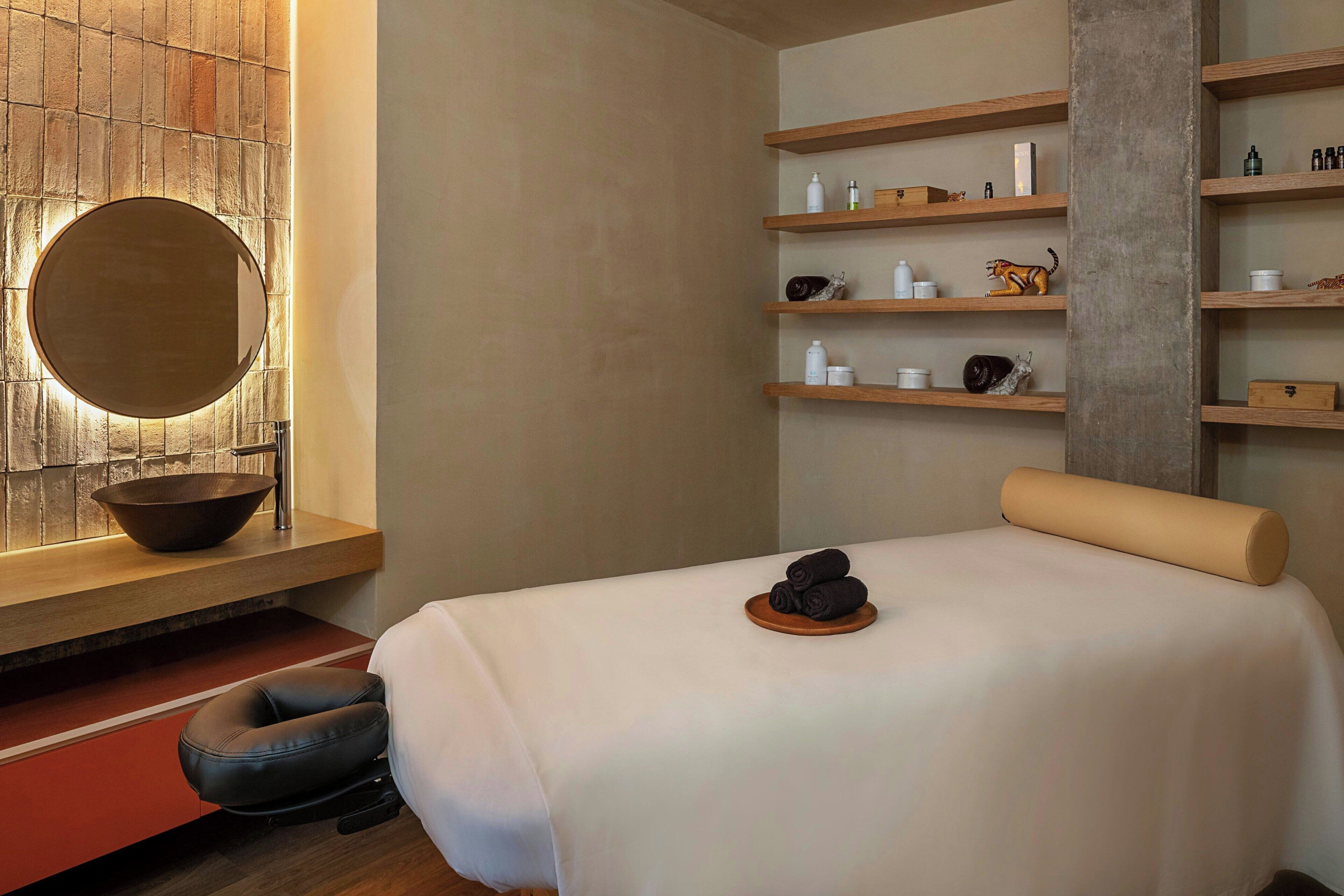 Une salle de spa moderne avec un grand miroir rond, des étagères en bois où sont rangés les produits de toilette, un fauteuil de massage et un lit avec une serviette enroulée.