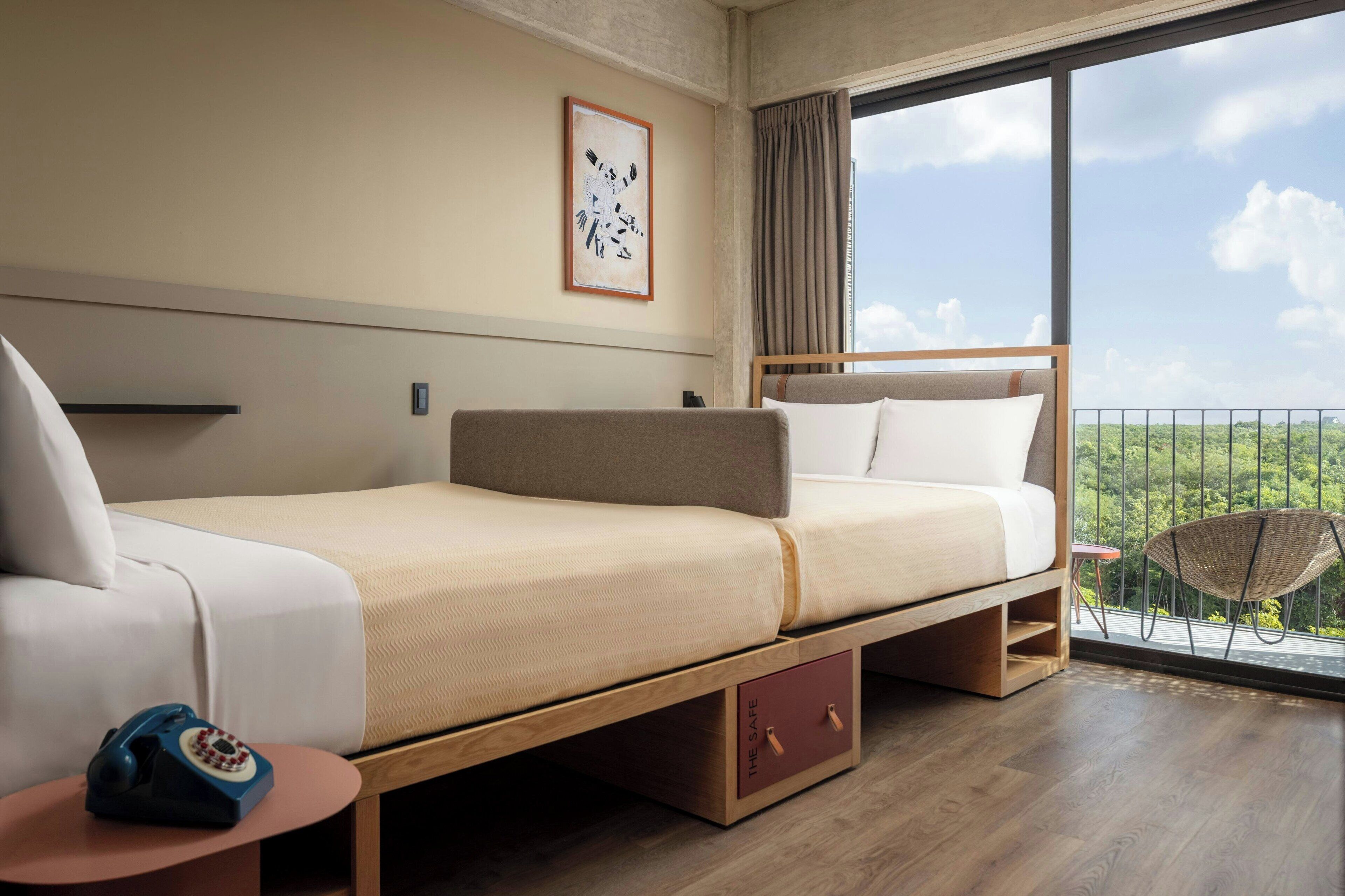 A modern hotel room with a bed, a small table with a telephone, a chair, and a balcony with a view of the greenery.