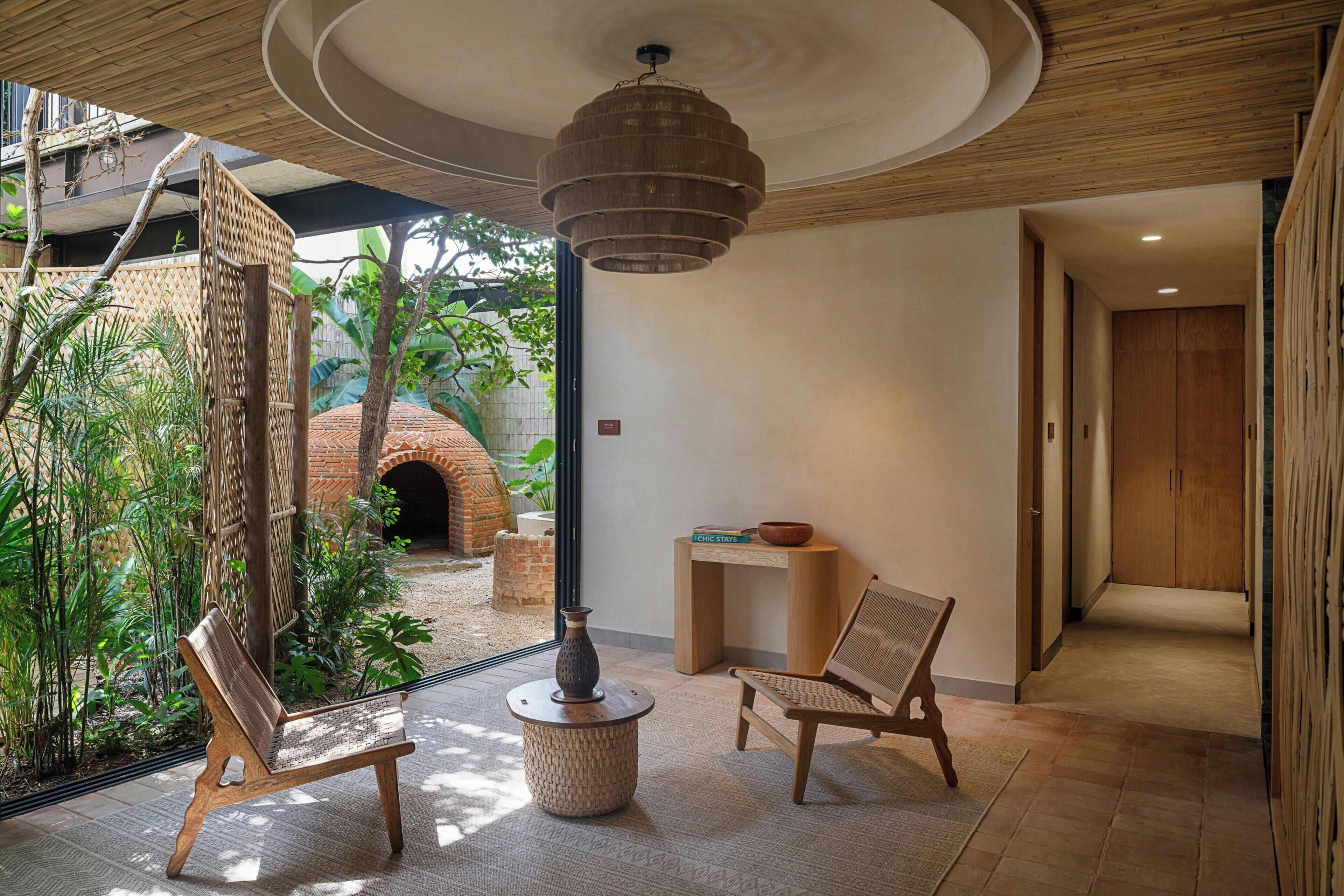 Une pièce avec un plafond en bois, un luminaire suspendu, un banc, une petite table et une vue sur un jardin extérieur avec un four en briques.