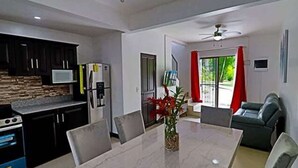 Una cocina moderna con armarios oscuros, un microondas y un refrigerador. Hay un comedor con una mesa de mármol y sillas, y una sala de estar con un sofá y una cortina roja.