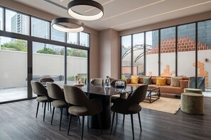 Un espace de repas moderne avec une table et des chaises en verre, un canapé avec des coussins et de grandes fenêtres offrant une vue sur les bâtiments et la verdure.