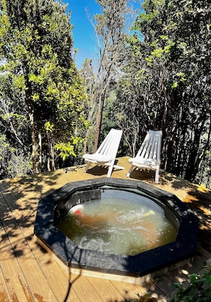 A hot tub surrounded by wooden deck and two white chairs, set in a wooded area.