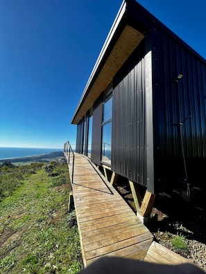 A modern building with a wooden deck leading to a large window, overlooking a coastal landscape.