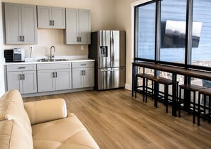 Une cuisine moderne avec des meubles gris, un réfrigérateur en acier inoxydable et une machine à café. On y trouve une table à manger en bois, entourée de tabourets, près d’une grande fenêtre.