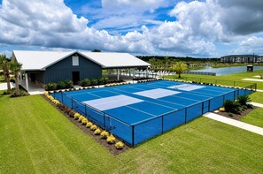 Un court de tennis entouré d’une clôture, doté d’un espace de repos couvert et bordé d’un bâtiment dont le toit est en pente.