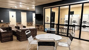 A clean, well-lit indoor area with wicker furniture, a round table, and a view of a laundry facility through large windows.