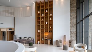 A modern living room with a curved white table, a sofa, and multiple seating options. There is a tall wooden shelving unit with various decorative items. - Séjour chic avec accès au spa + piscine (Baillargues)