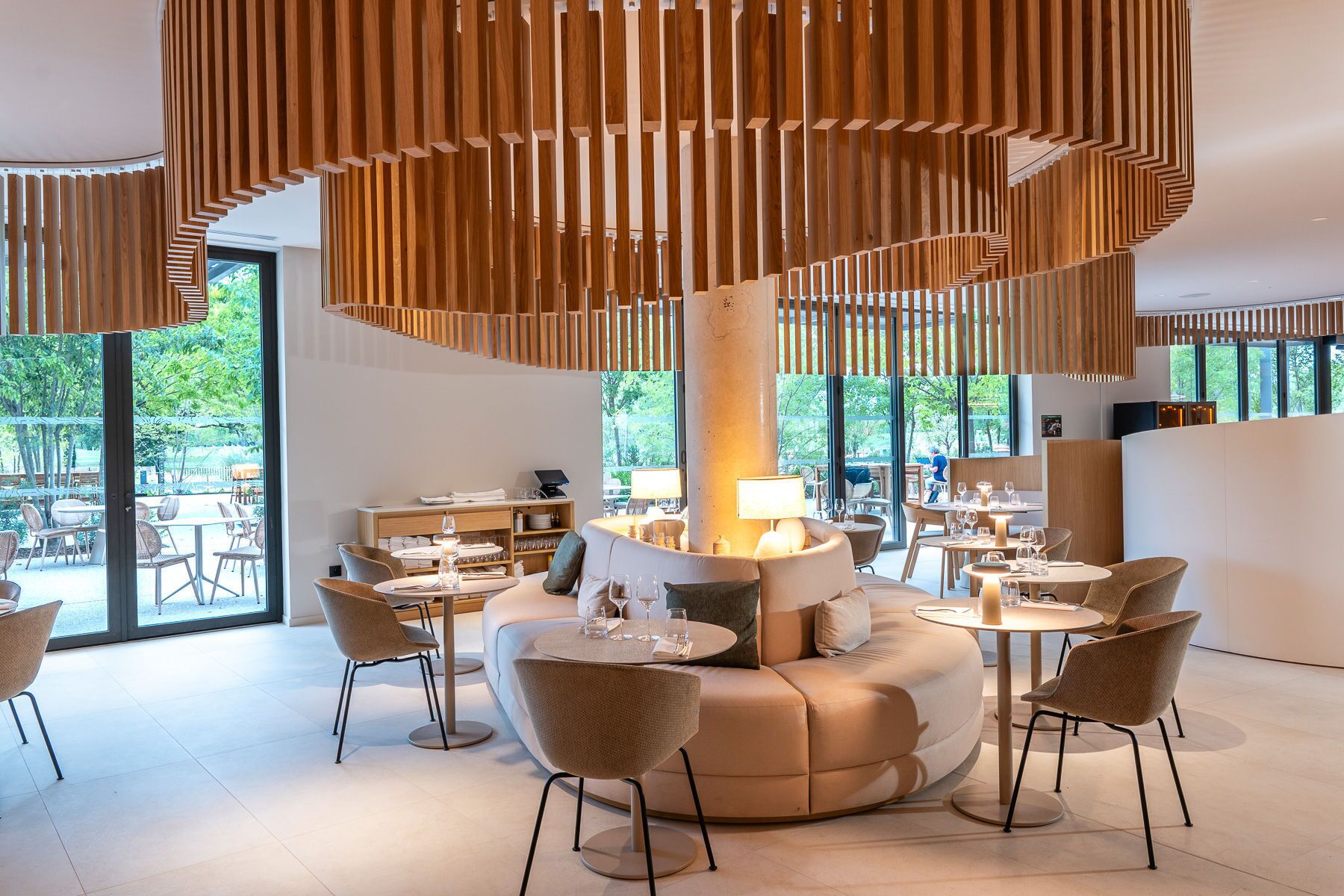 Een modern interieur met een centraal zithoek, meerdere tafels en een groot raam met uitzicht op het groen.