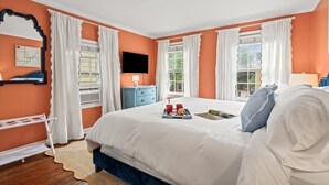 A bedroom with a large bed, two windows with white curtains, a flat-screen TV, a blue dresser, and a small table with a tray of items.