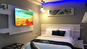 A modern bedroom with a large bed, a flat-screen TV mounted on the wall, a bedside table, and a small desk. - Madisonville Apartment  (Abuja)