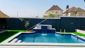 A swimming pool with a small waterfall feature, surrounded by a dark wall and a green lawn. - Madisonville Apartment  (Abuja)
