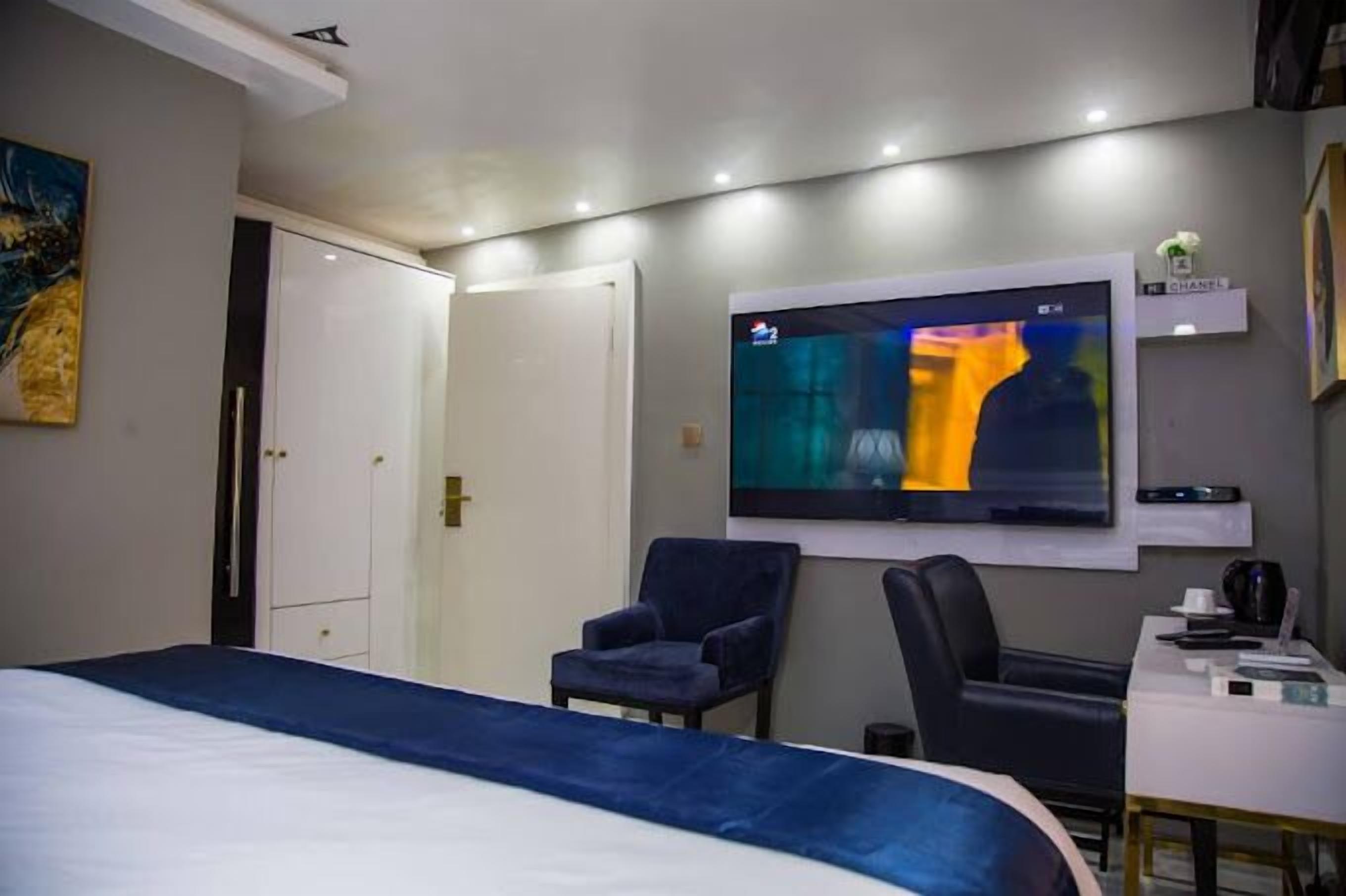 A hotel room with a bed, two chairs, a desk with a computer, and a TV mounted on the wall.
