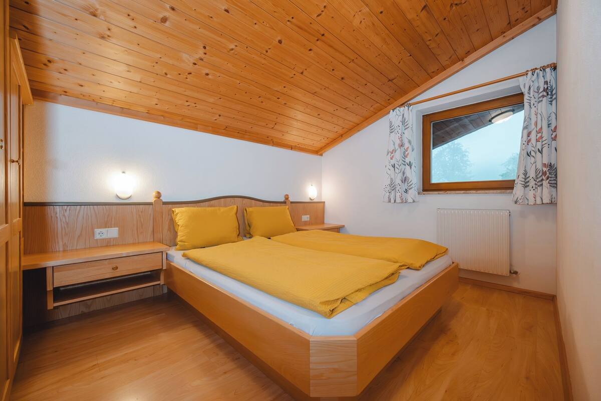 A bedroom with a wooden ceiling, a bed with yellow bedding, a nightstand, a window with curtains, and a radiator.