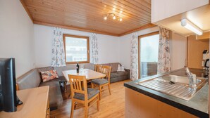 Un espace de vie compact doté d’un plafond en bois, d’un coin repas avec une table et des chaises, d’un canapé, d’une kitchenette avec un évier et de grandes fenêtres ornées de rideaux à motifs floraux.