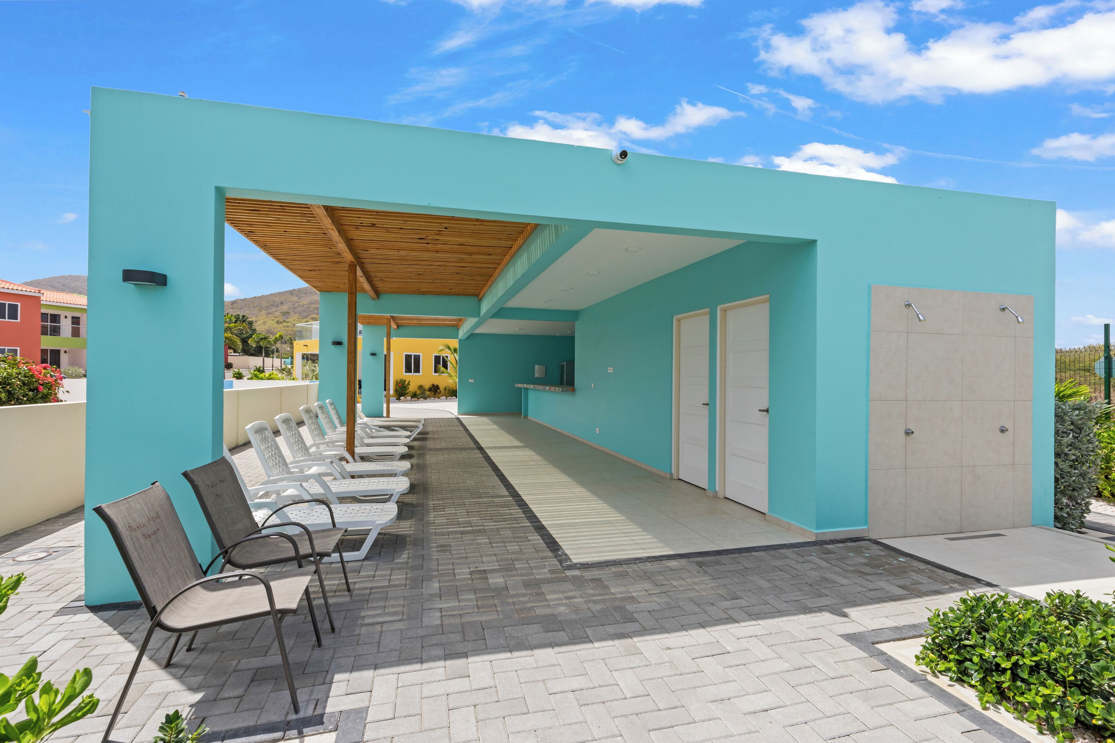 A covered outdoor seating area with lounge chairs and a wooden pergola, adjacent to a building with a light blue exterior and a tiled floor.