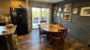 A dining area with a wooden table and chairs, a kitchen with a black refrigerator, and a balcony with a view of trees.