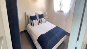 A bedroom with a bed, two pillows, a bedside table, and a window with curtains. - Apartment T2 - 40M² (40M²) (Saint-Martin-de-Seignanx)