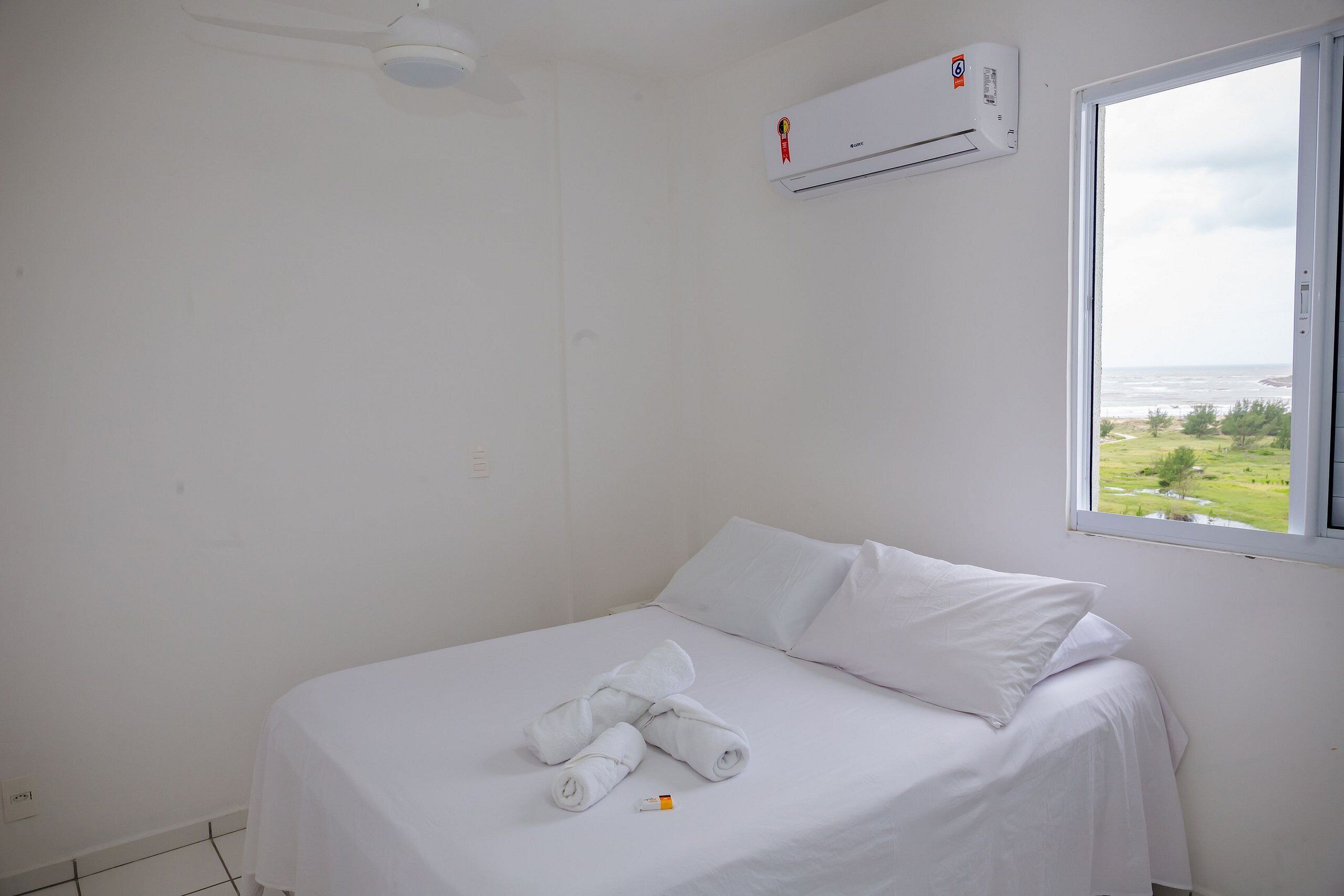A clean, white hotel room with a bed, a window with a view of the sea, and an air conditioning unit.