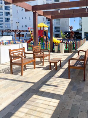 Een overdekt buitengebied met houten banken en tafels, een speelconstructie voor kinderen op de achtergrond en uitzicht op woongebouwen.