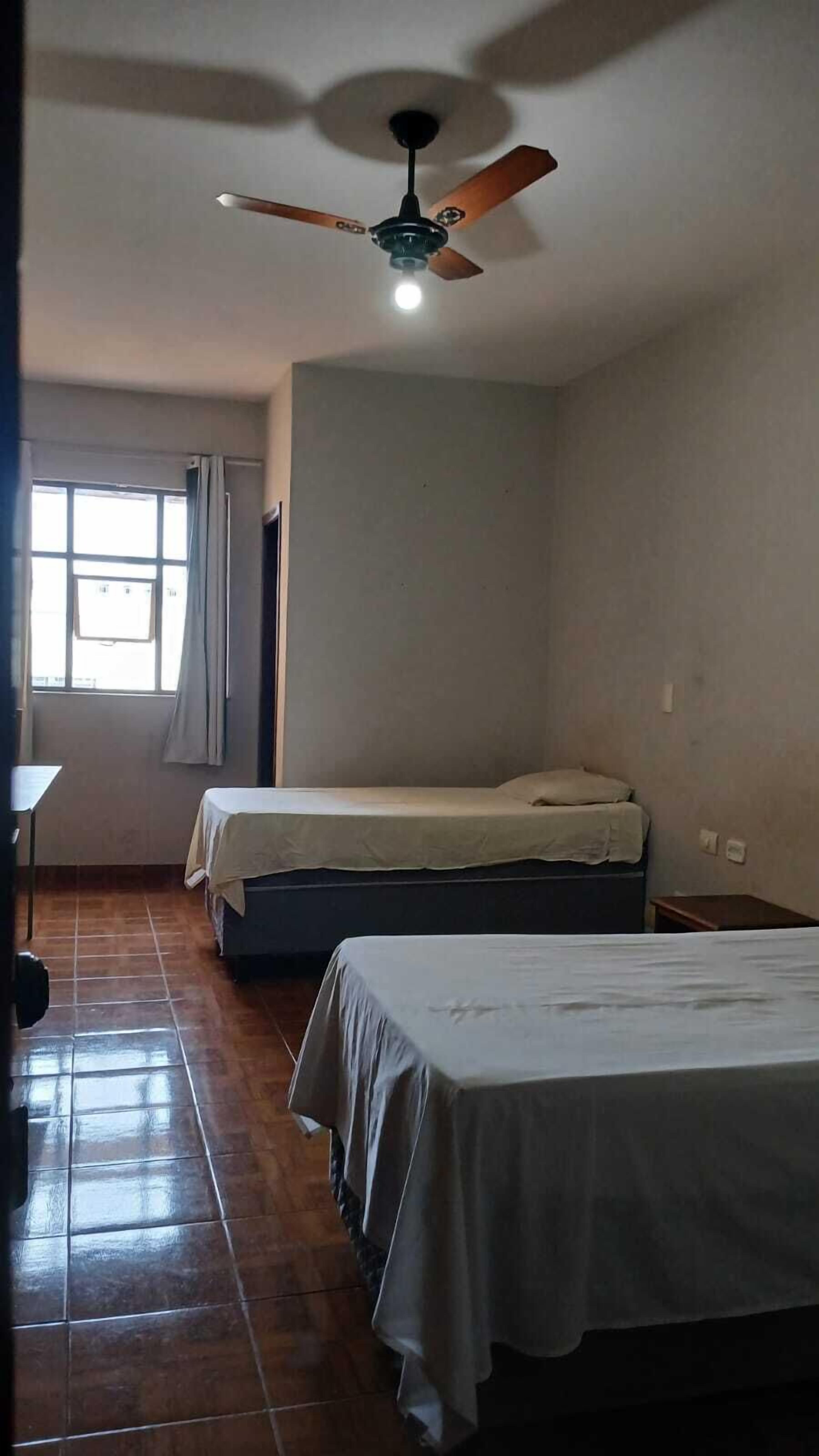 A room with two single beds, a ceiling fan, a window with curtains, and a tiled floor.