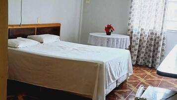 A hotel room with two beds, a small table with a vase of flowers, a desk with a computer, and patterned floor tiles.