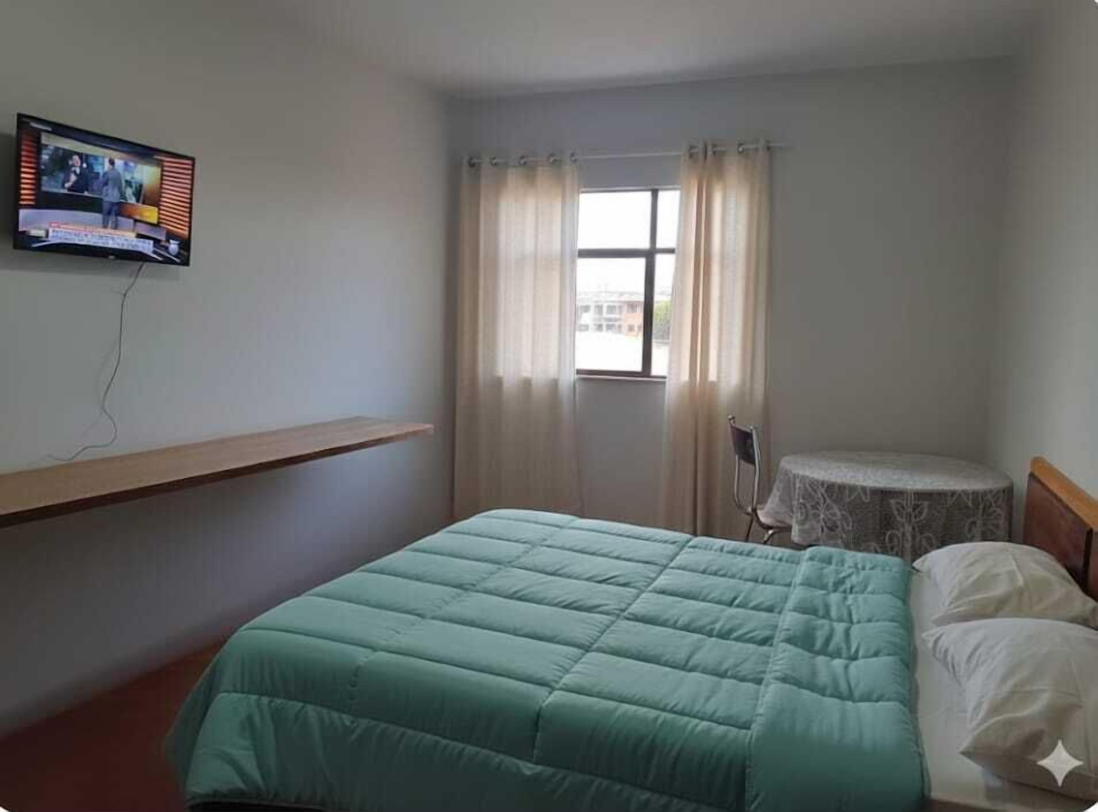A room with a bed, a small table, a TV mounted on the wall, and a window with curtains.