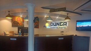 Ένας πάγκος υποδοχής στο θέρετρο DUNGA Lakeside, με μια πινακίδα που υποδεικνύει την περιοχή της υποδοχής, μια τηλεόραση τοποθετημένη στον τοίχο και έργα τέχνης πάνω από τον πάγκο.