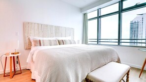 A modern bedroom with a large bed, a wooden headboard, a small round table with a lamp, and a view of buildings through large windows. - AKA Miami Brickell - Residence  (Miami)